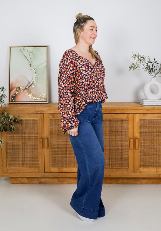 Woman wearing a patterned blouse and blue jeans standing in a room with wooden cabinets and decorative items.