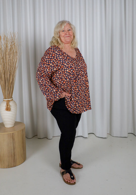 Woman wearing a patterned blouse and black pants standing in front of white curtains.