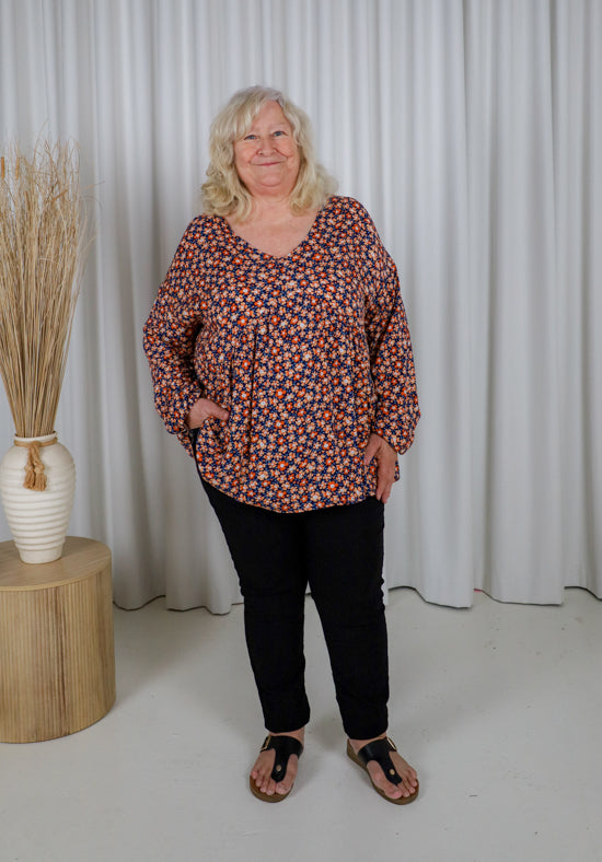 Woman wearing a patterned blouse and black pants standing in front of white curtains.