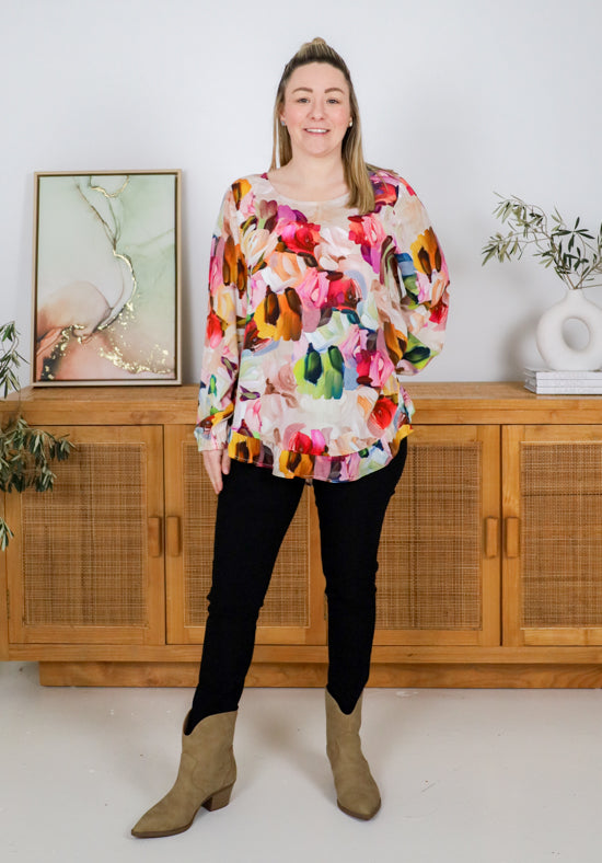 Woman wearing a colorful floral blouse standing in a room with wooden cabinets and decorative items.