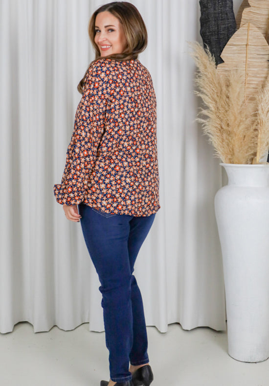 Woman wearing a patterned blouse and blue jeans standing in front of a white curtain.