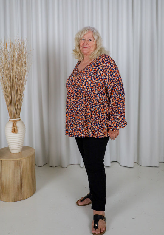 Woman wearing a patterned blouse and black pants standing in front of white curtains.