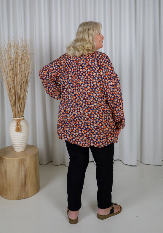 Woman wearing a patterned blouse standing in front of white curtains with a vase and decorative elements.