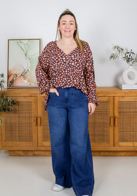 Woman wearing a floral blouse and blue jeans standing in a room with wooden furniture and plants.