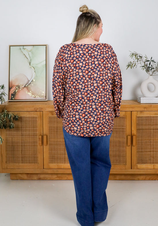 Person wearing a patterned blouse and blue jeans standing in front of a wooden cabinet.