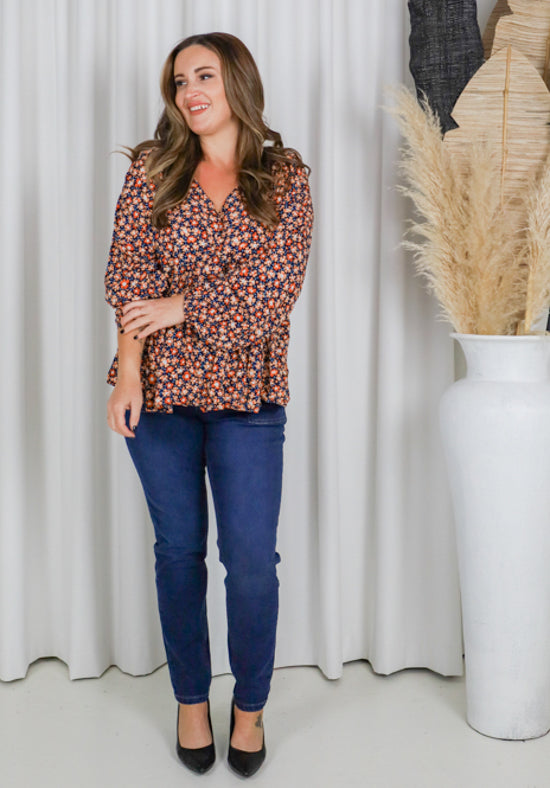 Woman wearing a patterned blouse and blue jeans standing in front of a white curtain.