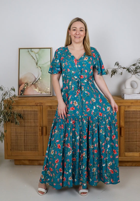 Woman wearing a teal floral dress standing in a room with wooden furniture and a painting.