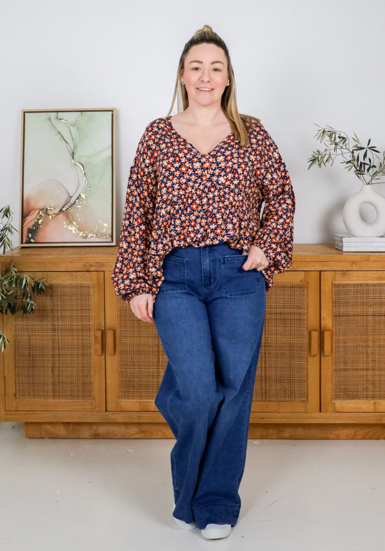 Woman wearing a floral blouse and blue jeans standing in a room with wooden cabinets and decorative items.