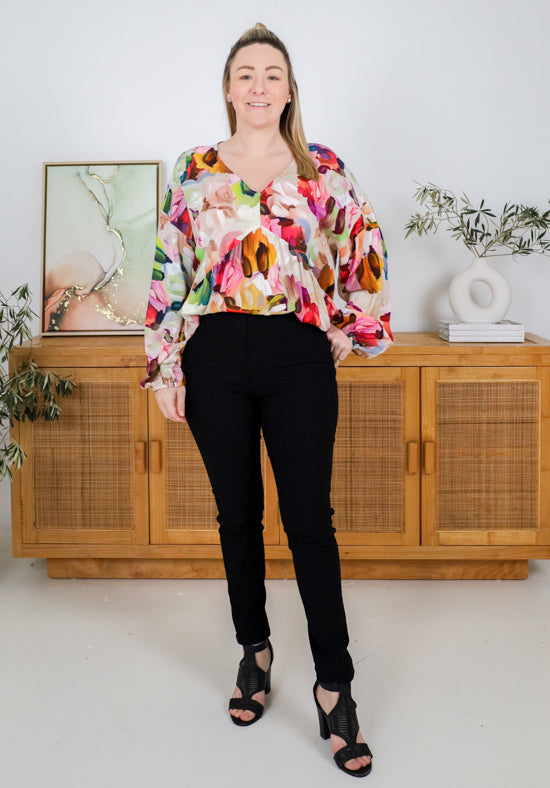 Woman wearing a colorful floral blouse and black pants standing in a room with wooden furniture and plants.