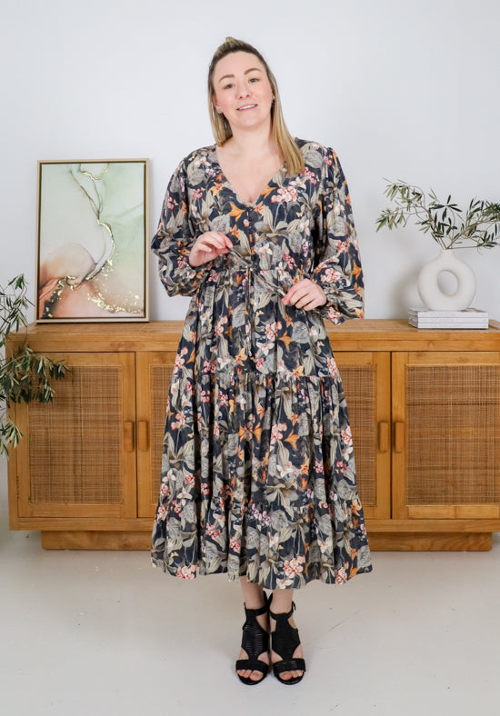 Woman wearing a floral dress standing in a room with wooden furniture and plants.
