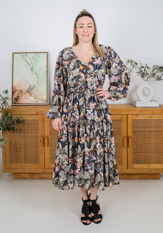 Woman wearing a floral dress standing in a room with wooden cabinets and plants.