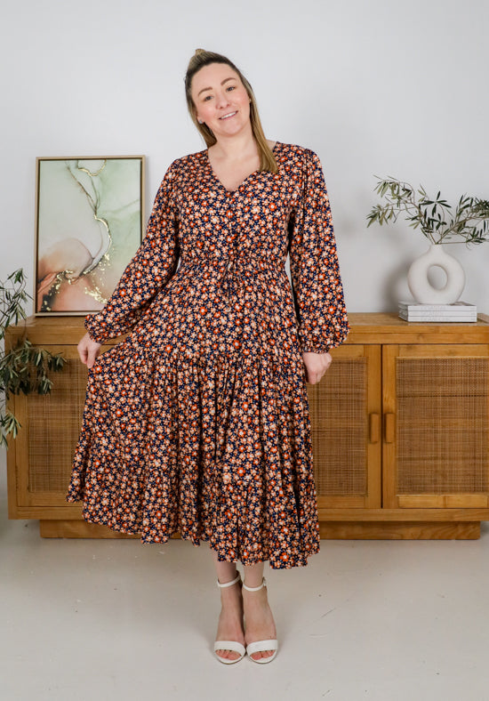 Woman wearing a floral dress standing in a room with wooden furniture and plants.