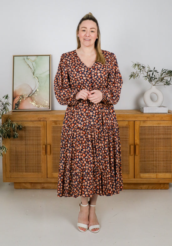 Woman wearing a floral dress standing in a room with wooden furniture and plants.