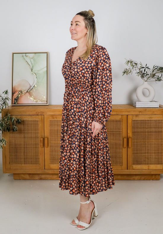 Woman in a floral dress standing in a room with wooden furniture and plants.