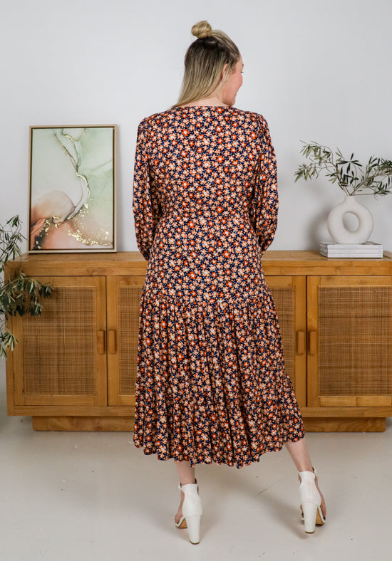 Woman wearing a floral dress standing in a room with wooden furniture and plants.