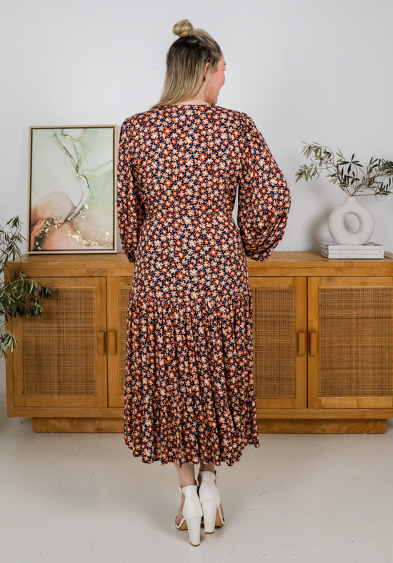 Woman wearing a floral dress standing in front of a wooden cabinet with plants.