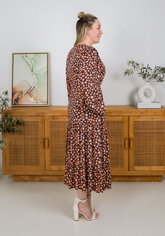Woman in a floral dress standing in a room with wooden furniture and plants.