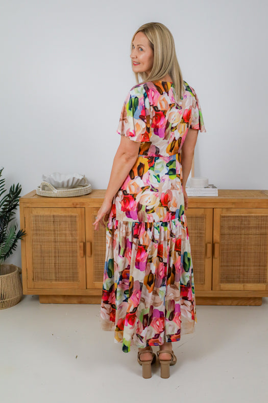 Woman wearing a colorful floral dress standing in a room with wooden furniture.