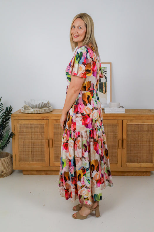 Woman wearing a colorful floral dress standing in a room with wooden furniture and decor.