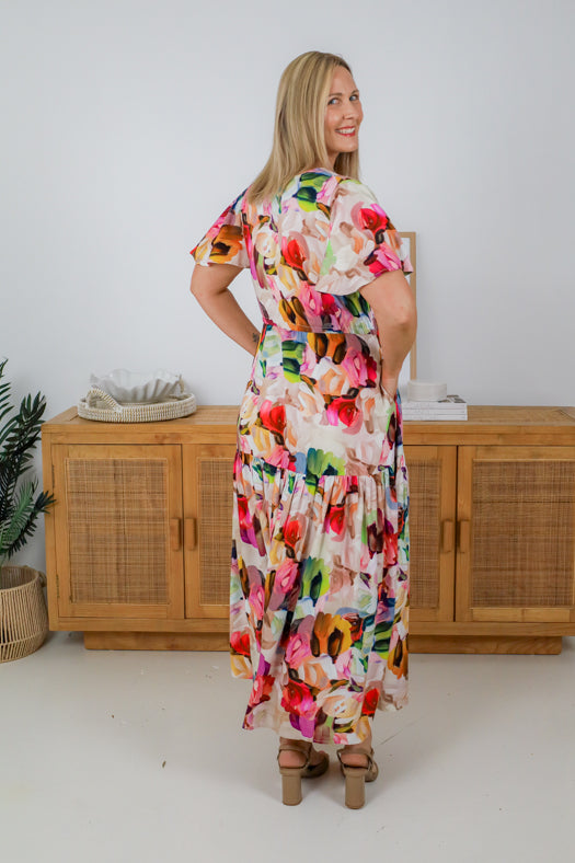 Woman wearing a colorful floral dress standing in a room with wooden furniture.
