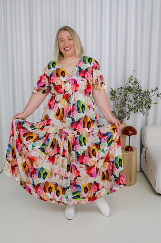 Woman wearing a colorful floral dress in a room with white curtains and a plant.