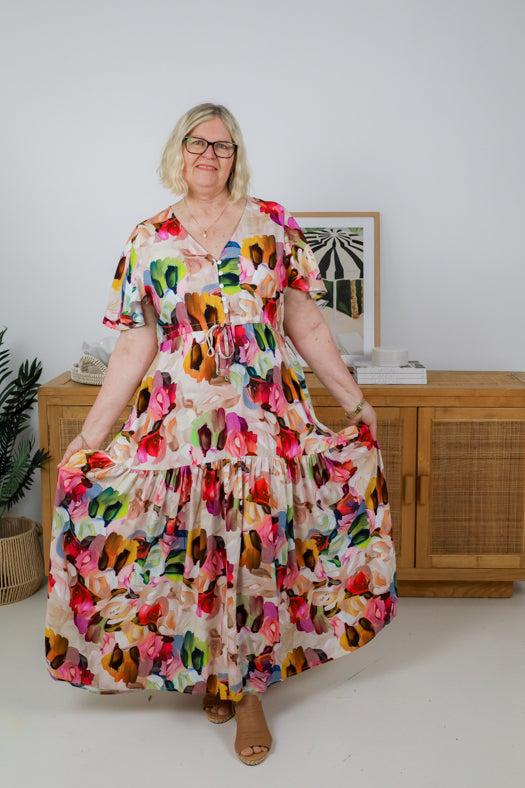Woman wearing a colorful floral dress in a room with a wooden cabinet and plant.