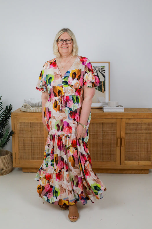 Woman wearing a colorful floral dress standing in a room with a wooden cabinet and framed picture.