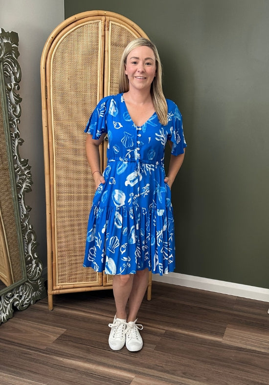 Woman in a blue dress standing in a room with a wicker cabinet and decorative mirror.