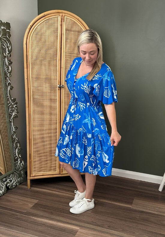 Woman wearing a blue dress with white patterns standing in a room with a wicker cabinet and decorative mirror.