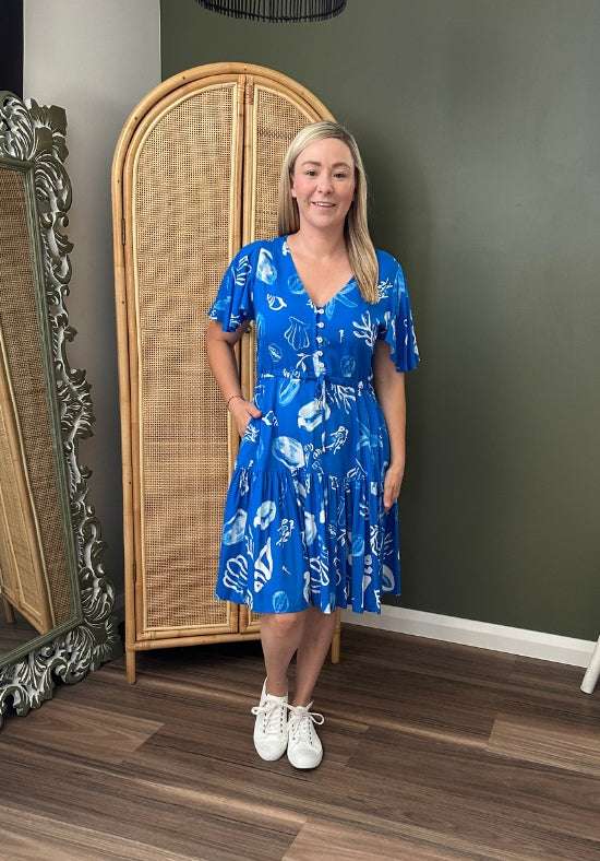 Woman in a blue dress standing in a room with a rattan cabinet and decorative mirrors.