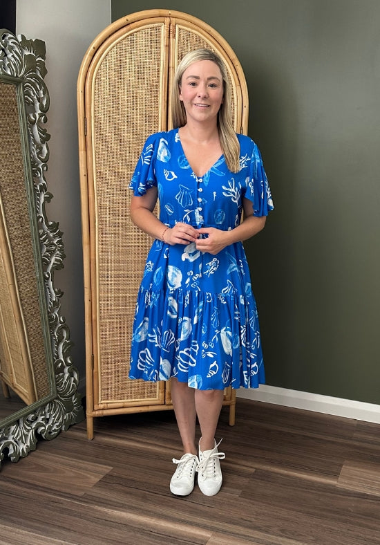Woman wearing a blue dress with a white pattern standing in a room with a wicker cabinet and decorative mirror.