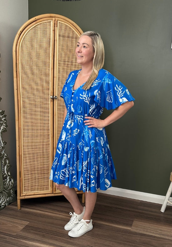 Woman wearing a blue dress with white patterns standing in a room with a wicker wardrobe.