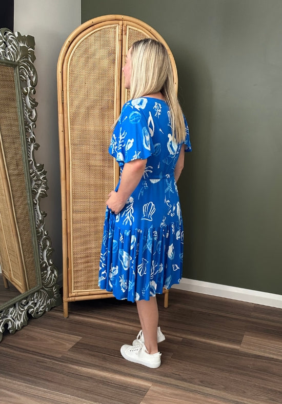 Woman wearing a blue dress with white patterns standing in front of a rattan mirror.