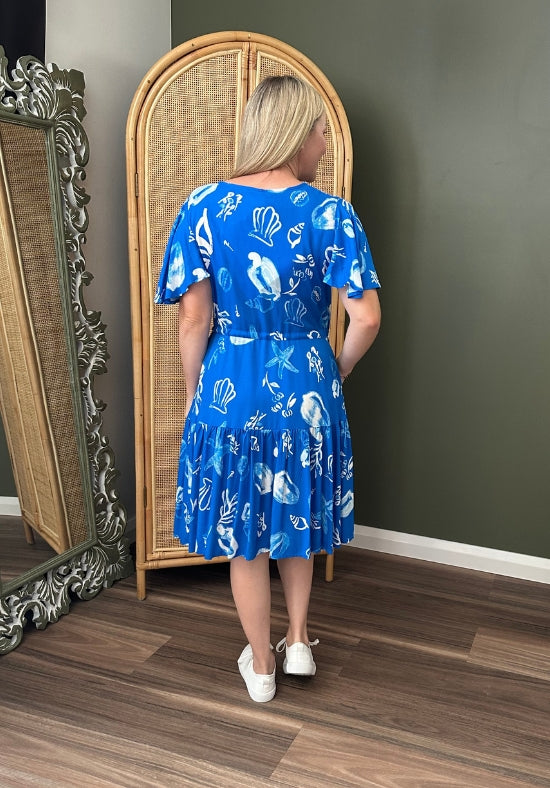 Woman wearing a blue dress with white patterns standing in front of a mirror.