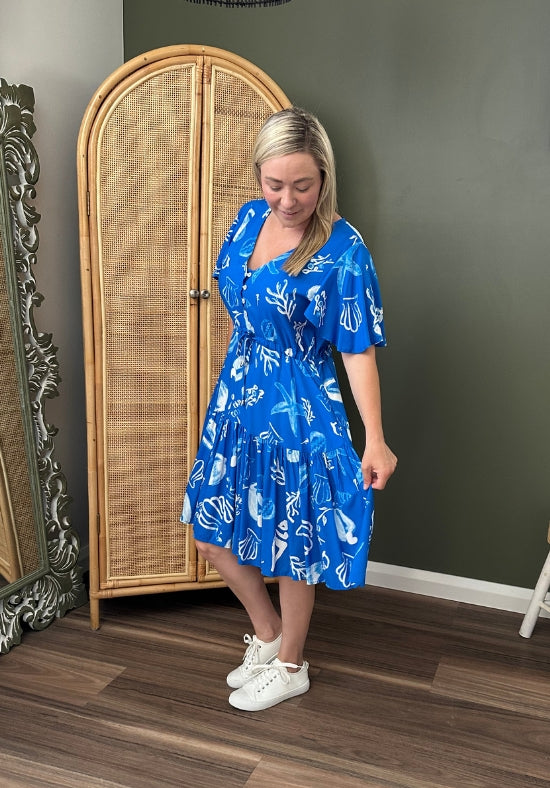Woman wearing a blue dress with a white pattern in a room with a wooden wardrobe and decorative mirror.
