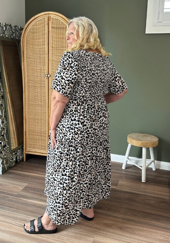 Woman wearing a leopard print dress standing in a room with a wicker cabinet and wooden floor.