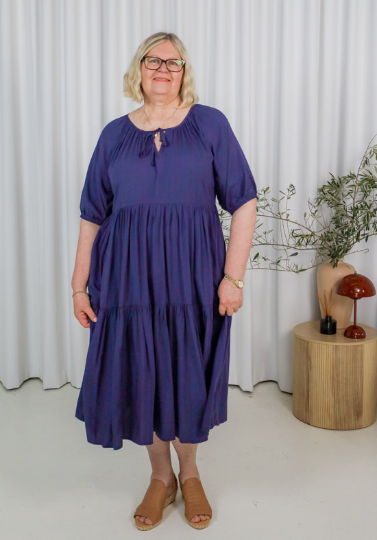 Woman wearing a blue dress standing in a room with a white curtain and wooden table.