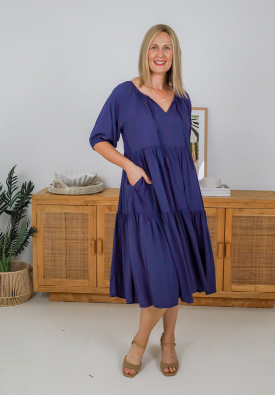 Woman wearing a blue dress standing in a room with wooden furniture and decor.