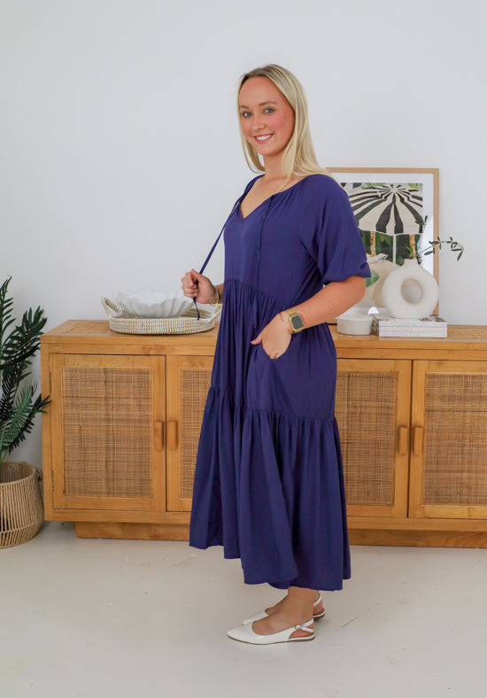Woman wearing a purple dress standing in a room with wooden furniture and decor.