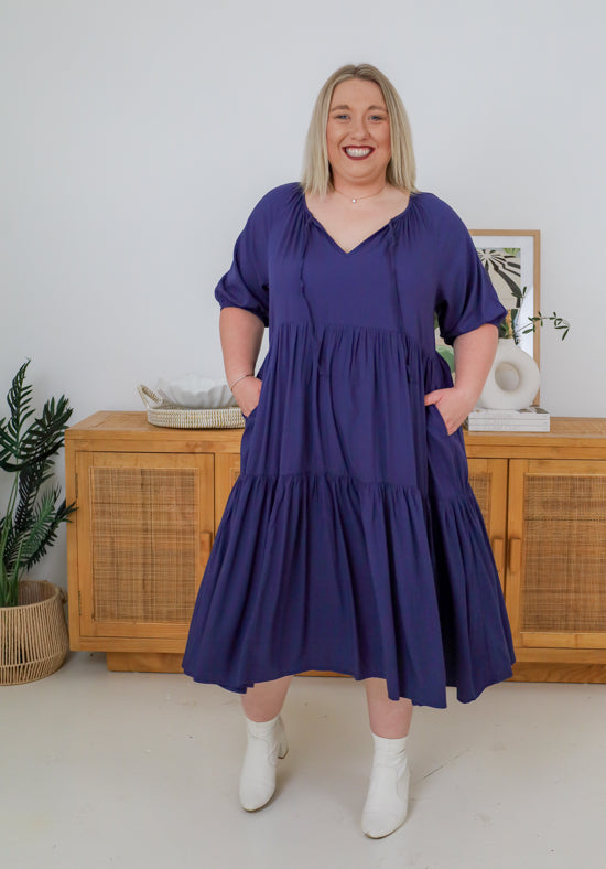 Woman wearing a purple dress standing in a room with wooden furniture and decor.