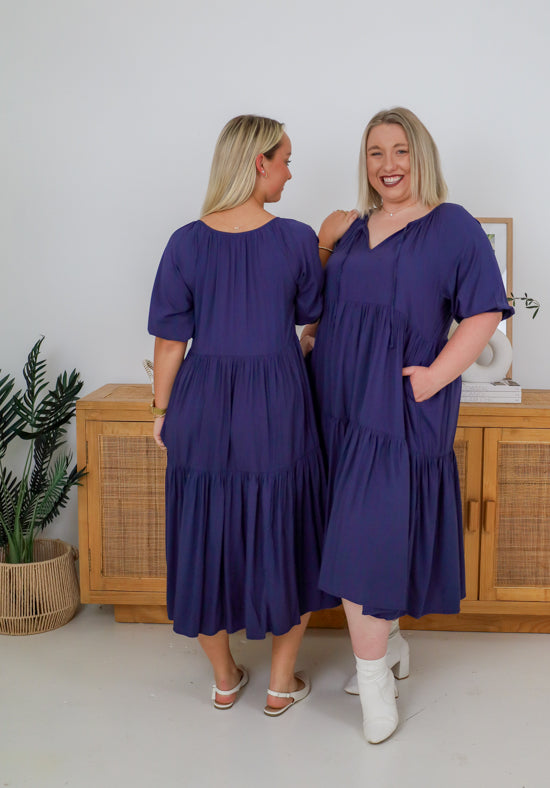 Two women wearing matching purple dresses standing in a room with a wooden cabinet and plant.