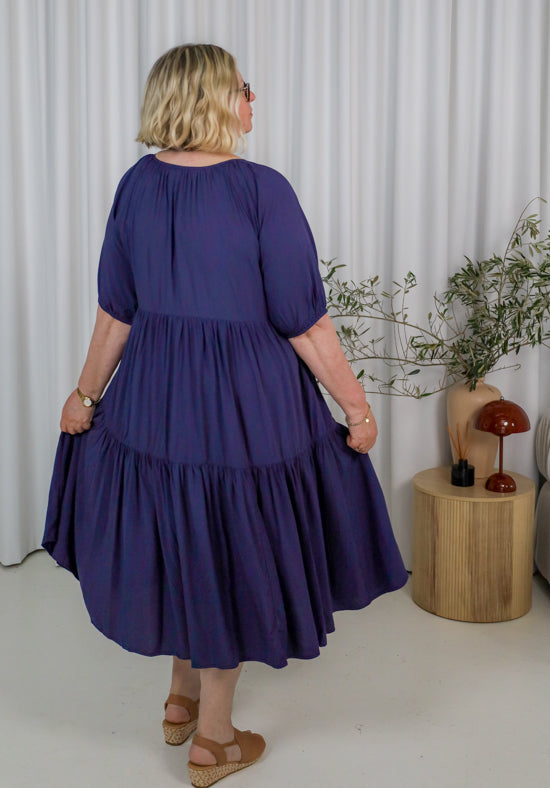 Woman wearing a purple dress standing in a room with a white curtain and wooden side table.
