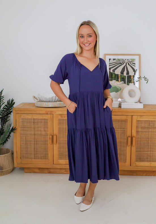 Woman wearing a blue dress standing in front of wooden cabinets.