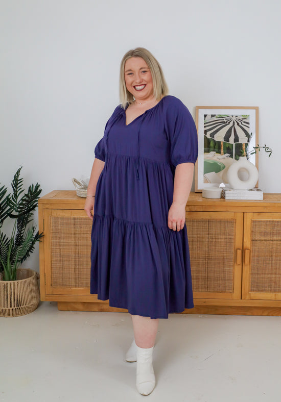 Woman wearing a purple dress standing in a room with wooden furniture and decor.