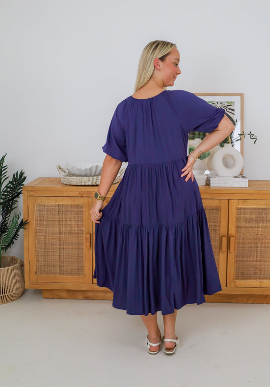 Woman wearing a purple dress standing in a room with wooden furniture and decor.