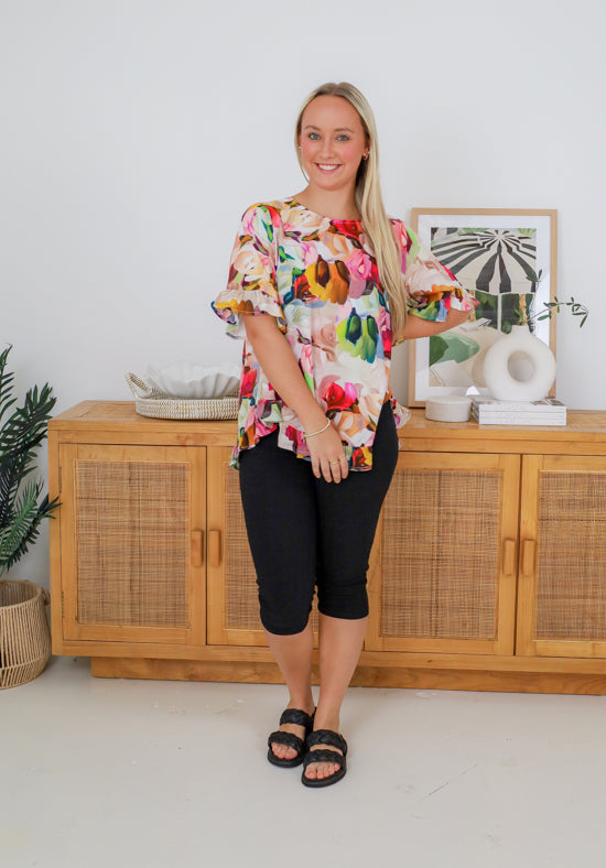 Woman wearing a colorful floral blouse and black shorts standing in front of a wooden cabinet.