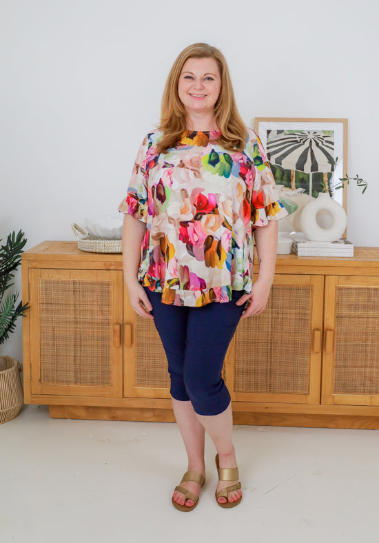 Woman wearing a colorful floral blouse and navy pants standing in a room with wooden furniture and decor.