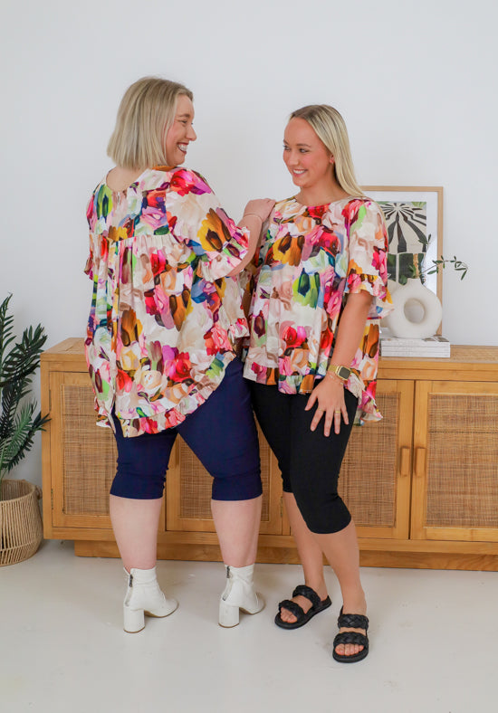 Two women wearing colorful floral tops and navy shorts standing in a room with a wooden cabinet and plant.