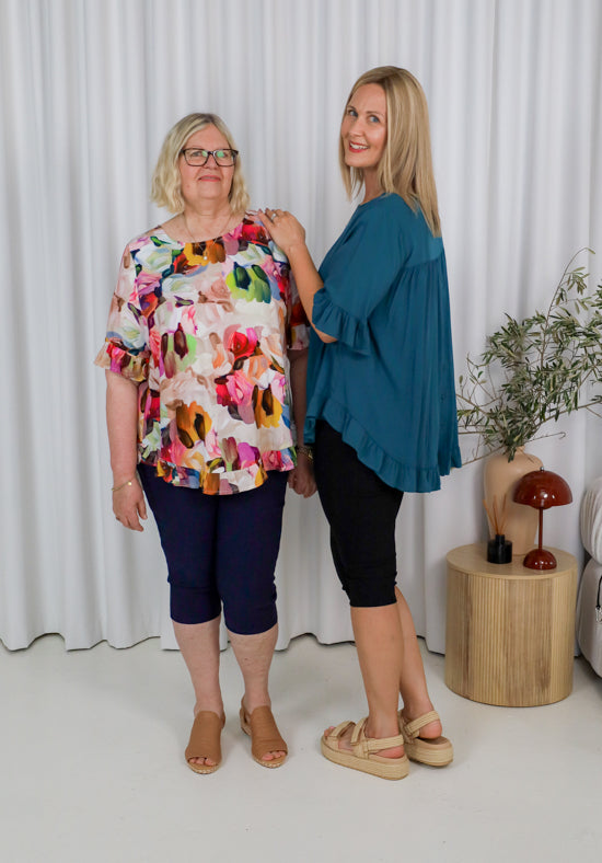 Two women standing next to each other wearing colorful tops and dark pants against a white curtain background.