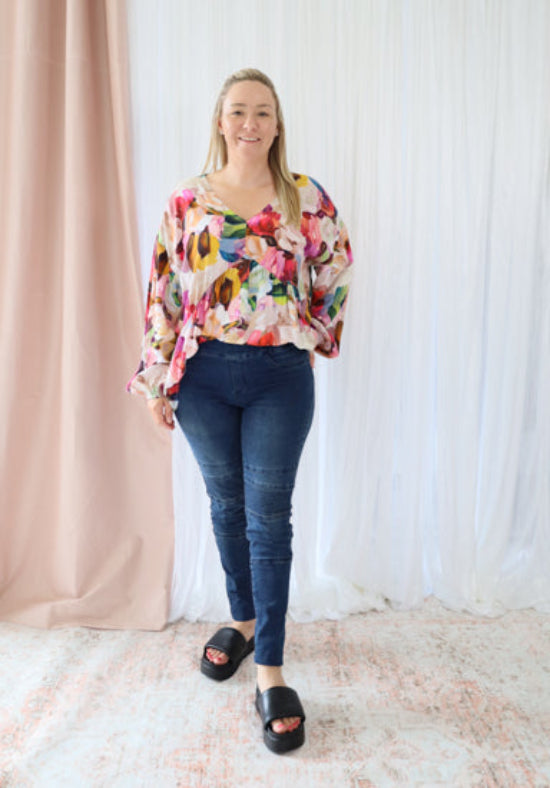 Woman wearing a colorful floral blouse and blue jeans standing against a white curtain background.
